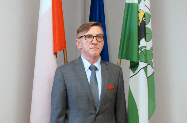 Mężczyzna stoi w sali urzędu. Za nim jest stojak z 3 flagami - Polski, Unii Europejskiej i Powiatu Oleckiego.