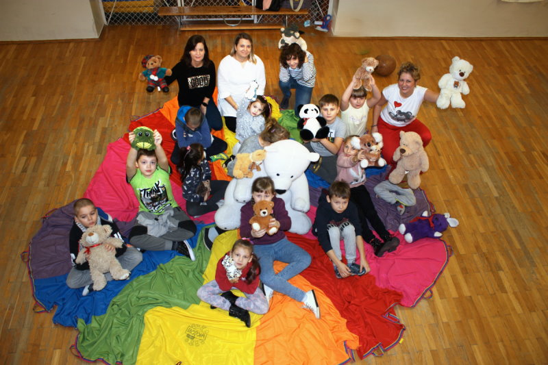 17 osób siedzi na kolorowym materiale na podłodze w sali. W ręku trzymają maskotki – misie.