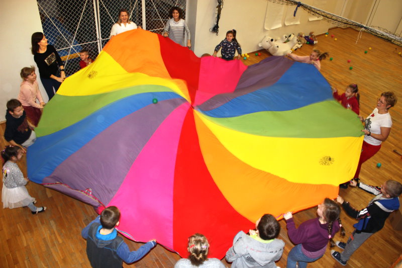 16 osób stoi w kółku w sali. W rękach trzymają wspólnie kolorowy materiał. Na podłodze leżą maskotki – misie i małe piłki.