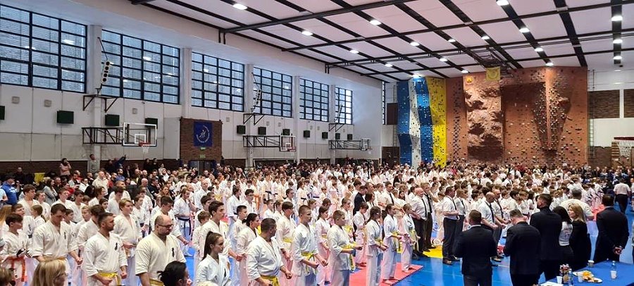 W sali sportowej stoi obok siebie kilkadziesiąt osób. Niektórzy z nich są ubrani w kimono; są zawodnikami karate.