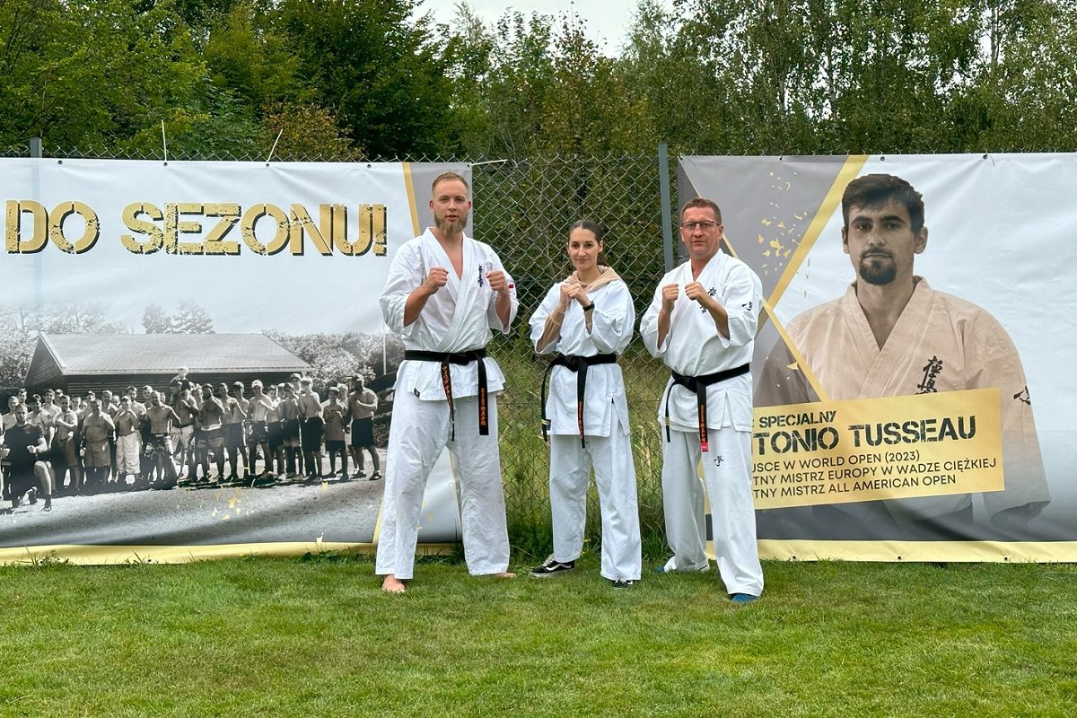 Na podwórku stoi obok siebie kobieta i 2 mężczyzn. Są oni ubrani w kimono do karate. Za nimi ogrodzenie z banerami.