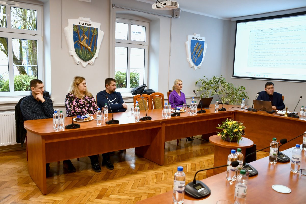 W pomieszczeniu w budynku przy stole siedzi 3 mężczyzn i 2 kobiety. Przed jednym z mężczyzn stoi laptop. Na stole są między innymi mikrofony, szklanki, butelki z wodą. Na środku stolik z kwiatami. W tle ściana z ekranem, oknami i herbami.