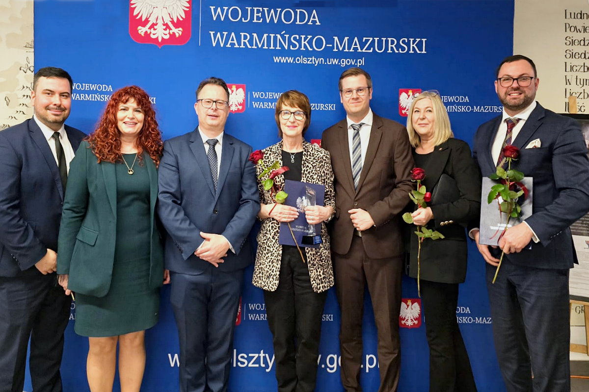 W pomieszczeniu obok siebie stoi 4 mężczyzn i 3 kobiety. Dwie z kobiet trzymają róże, a jedna z nich jeszcze statuetkę i teczkę z herbem. Jeden z mężczyzn trzyma różę i teczkę z herbem.