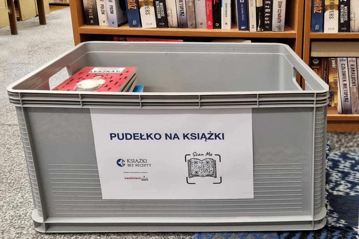 W pomieszczeniu na podłodze stoi pudło z książkami. Na pudle jest kartka, a na niej napisy. Pudło na książki i logo z napisem: książki bez recepty. Obok kod QR z napisem w języku angielskim: scan me. W tle regały z książkami.