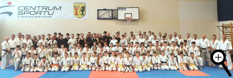 Duża grupa osób w strojach do karate i trenerzy pozuje wspólnie w sali sportowej. Na ścianie widoczny jest baner z napisem „Centrum Sportu w Gminie Lesznowola” i herbem gminy.