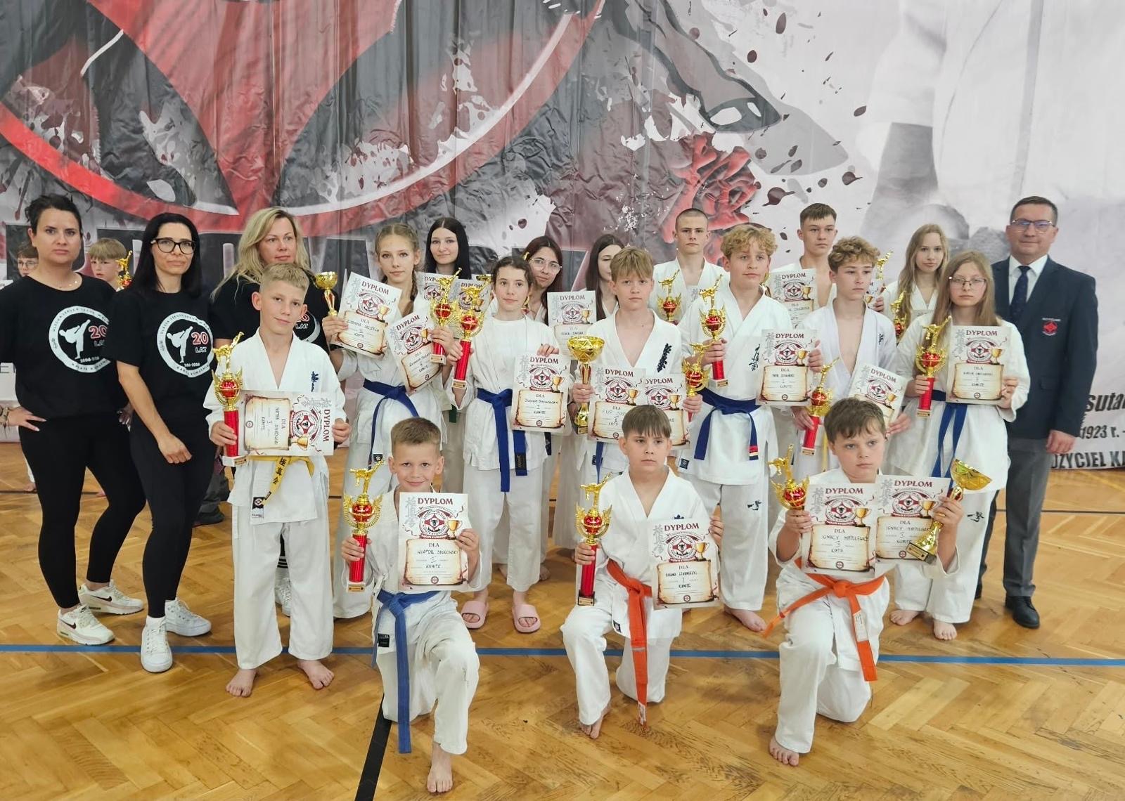 Grupa młodych zawodnik&oacute;w karate z Olecka pozuje do wsp&oacute;lnego zdjęcia w hali sportowej. Uczestnicy w białych kimonach prezentują puchary i dyplomy zdobyte podczas turnieju. Towarzyszą im trenerzy i opiekunowie.