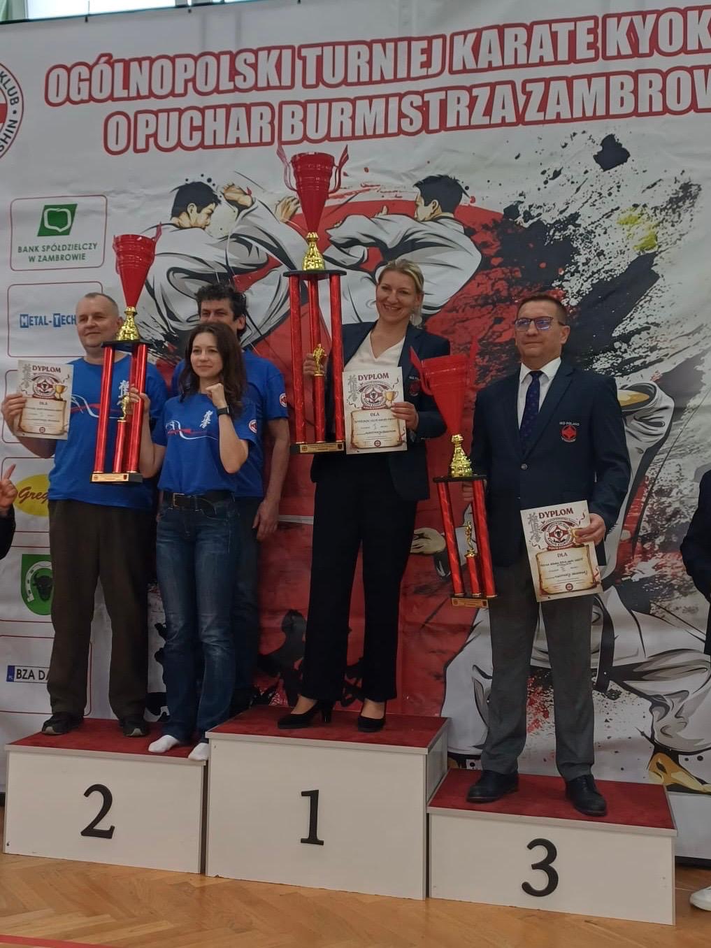Trenerzy i przedstawiciele klub&oacute;w karate stoją na podium podczas dekoracji drużynowej na Og&oacute;lnopolskim Turnieju Karate Kyokushin o Puchar Burmistrza Zambrowa. Uczestnicy prezentują puchary i dyplomy, a w tle widoczny jest baner zawod&oacute;w.