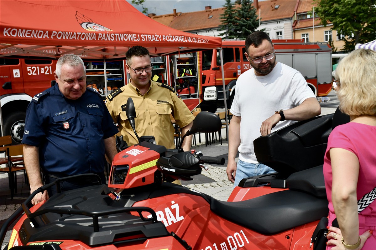 Odwiedzający oglądają czerwony quad lub pojazd ratowniczy prezentowany przez służby podczas festynu. Przy stoisku rozmawiają funkcjonariusze i uczestnicy wydarzenia.