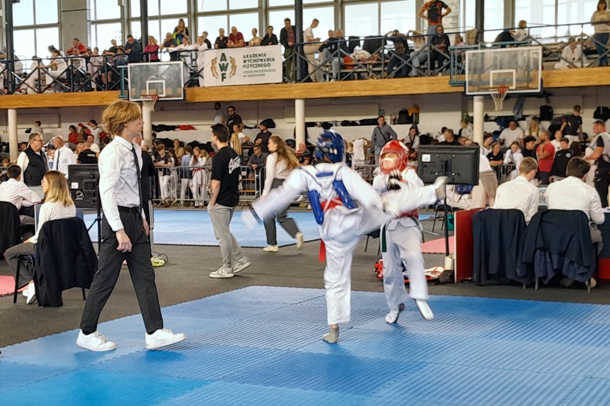 Zawodnicy podczas walki taekwondo na niebieskiej macie sportowej. W tle widoczni sędzia, trenerzy, publiczność na trybunach oraz stanowiska sędziowskie podczas zawod&oacute;w.